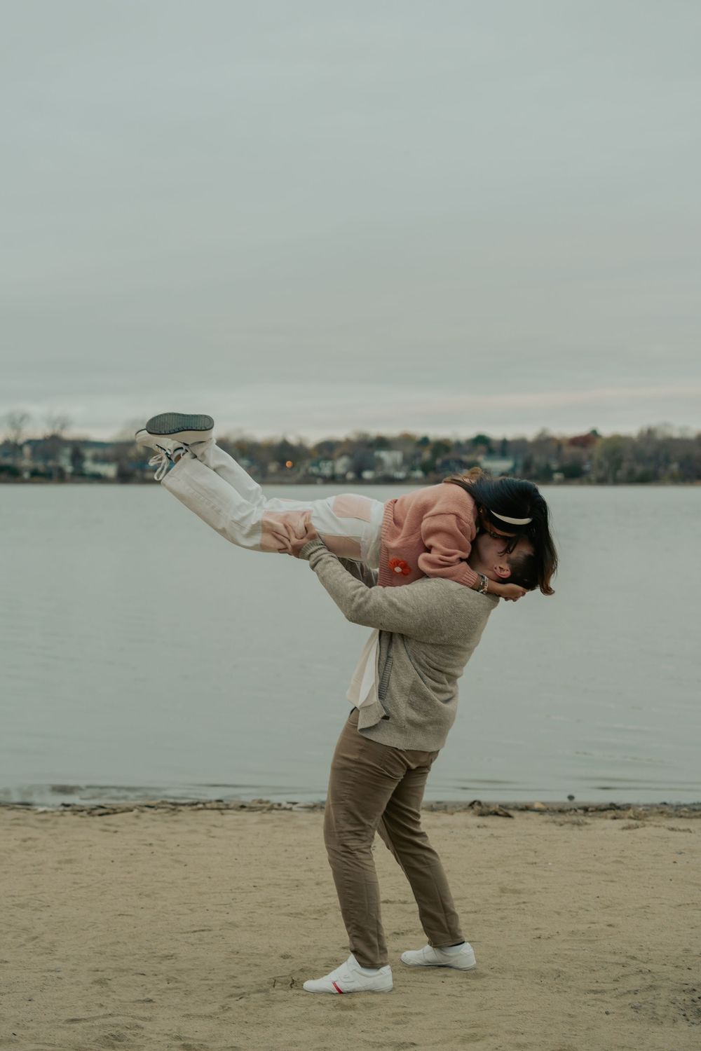 A person in beige clothing performs a graceful dance sequence on a sandy beach with a lake in the background.
