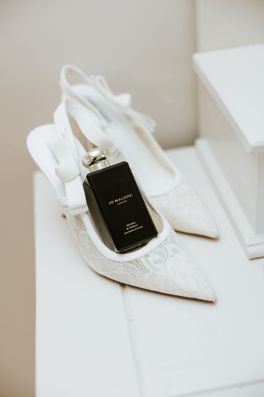 Elegant white lace slingback wedding heels with pointed toe displayed with a black Jo Malone perfume bottle. Castle Leslie