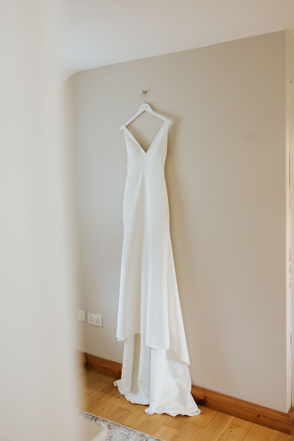 A simple white wedding dress hangs elegantly against a cream colored wall with wooden baseboards. Castle Leslie