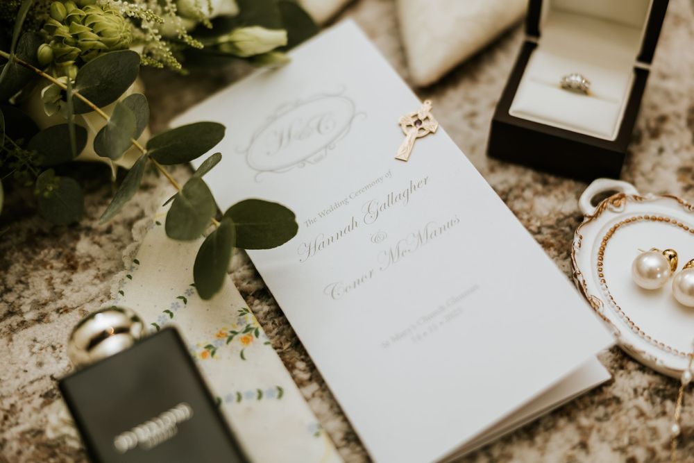 Wedding details including white invitations, eucalyptus leaves, and delicate gold jewelry arranged on a textured surface. Castle Leslie