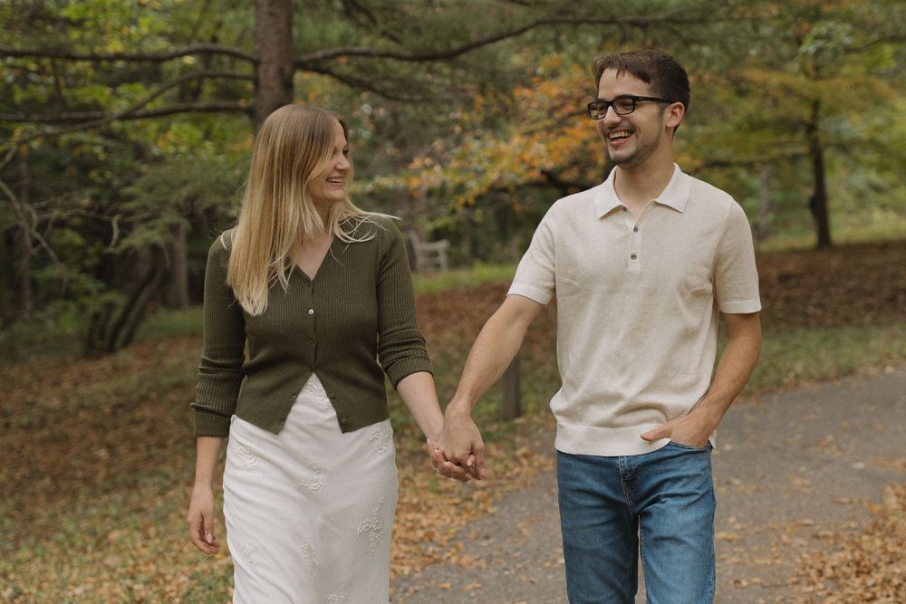 A couple walks hand in hand along a path covered in fallen autumn leaves.
