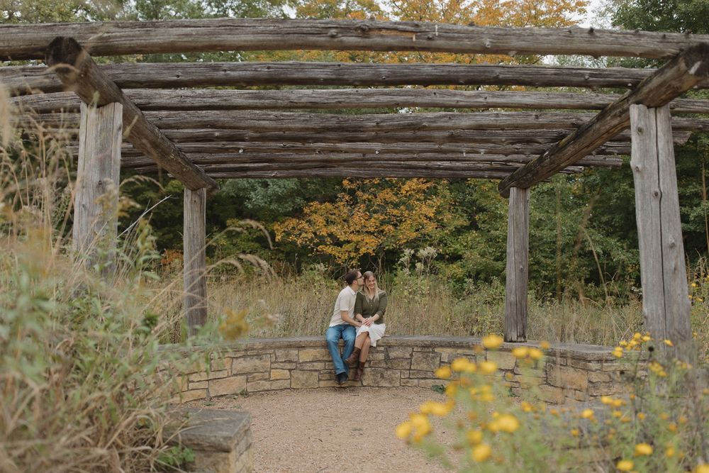 A couple sits together under a rustic wooden pergola surrounded by fall flowers.