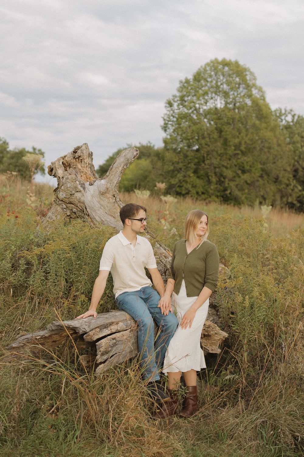 A couple sits together on a fallen log in a golden wildflower meadow during sunset.