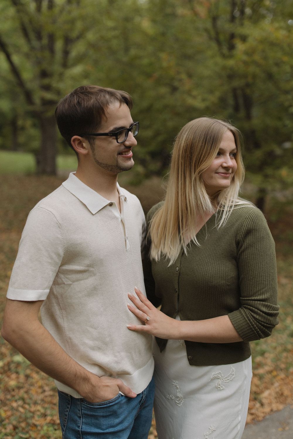 A couple smiles together on a fall day surrounded by trees with changing leaves.