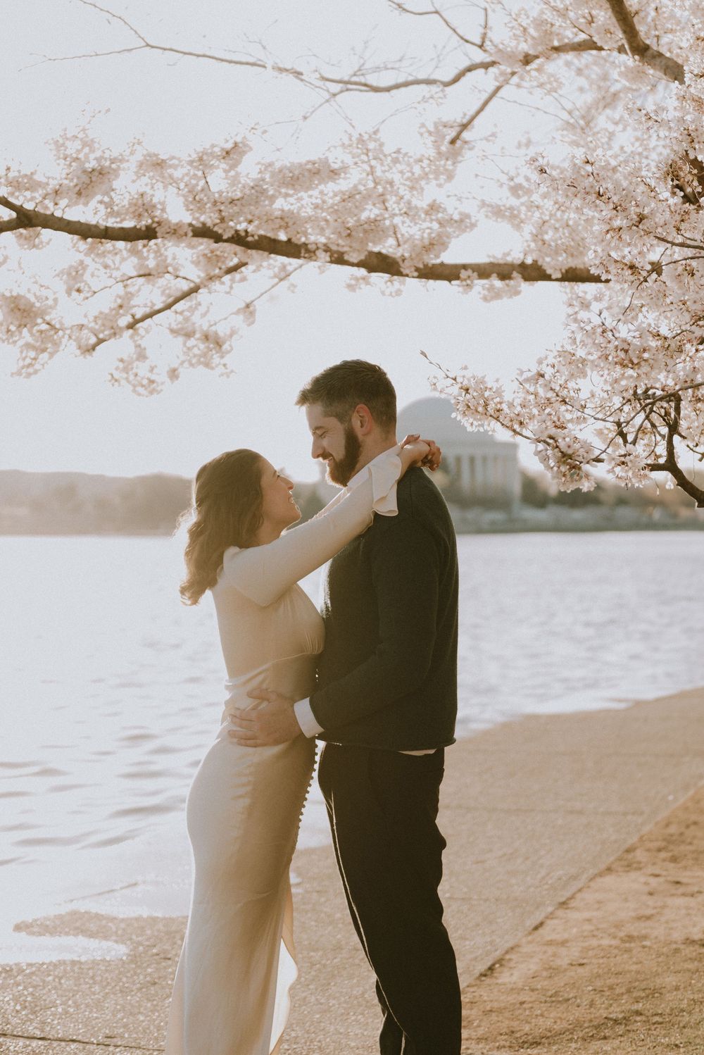 Romantic couple embraces under blooming cherry blossoms by the water during sunset at the Jefferson Memorial.