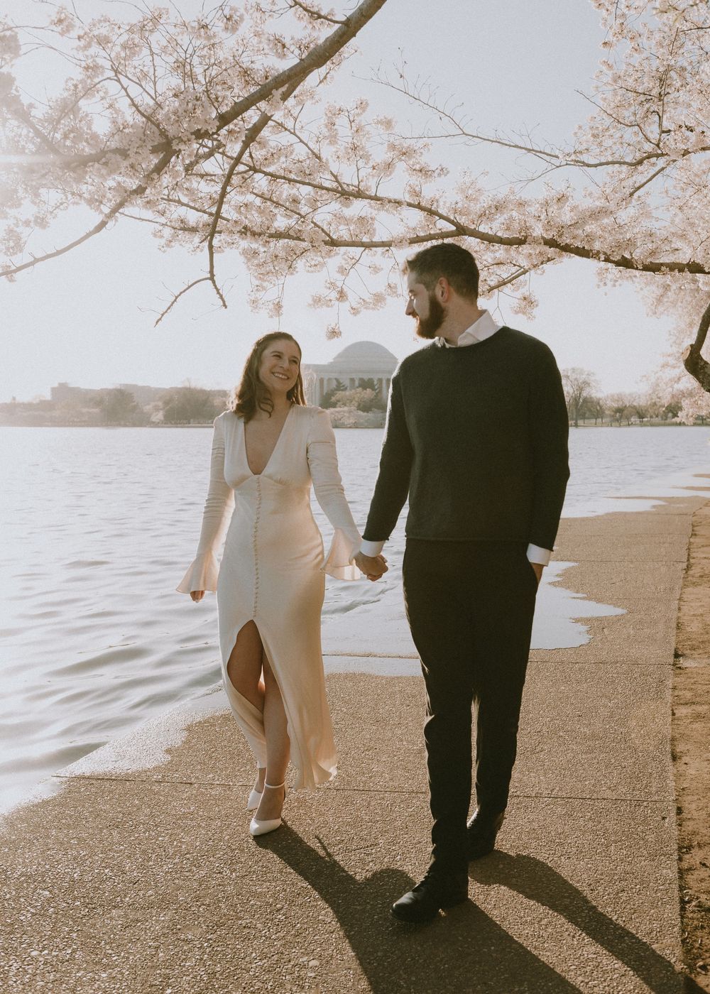 A couple strolls along the waterfront beneath blooming cherry blossom trees at sunset.