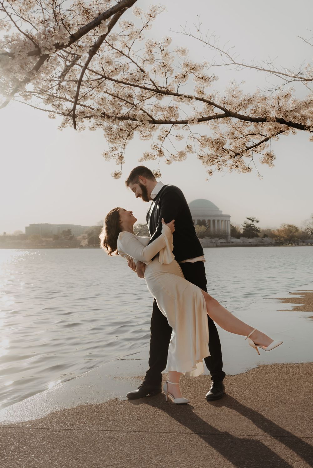 A couple shares an intimate dance moment by the water under blooming cherry blossom branches.
