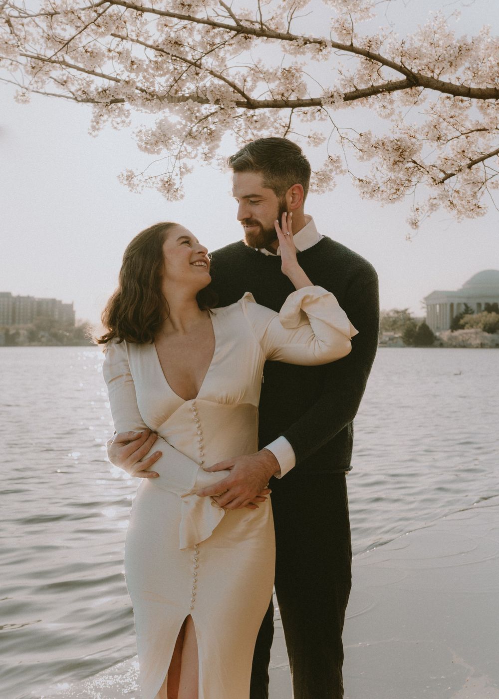 A romantic couple shares tender moments under cherry blossoms by the water during golden hour.