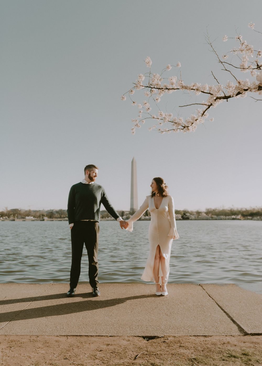 A couple walks along the Tidal Basin with cherry blossoms and Washington Monument in view during sunset.