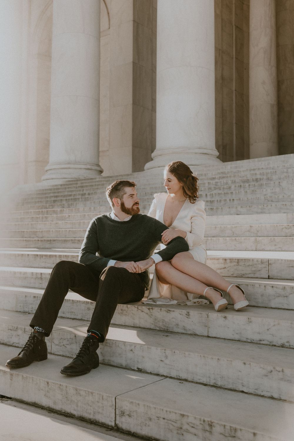 Stylish couple sits on marble steps of a classical building with grand columns in warm sunlight.