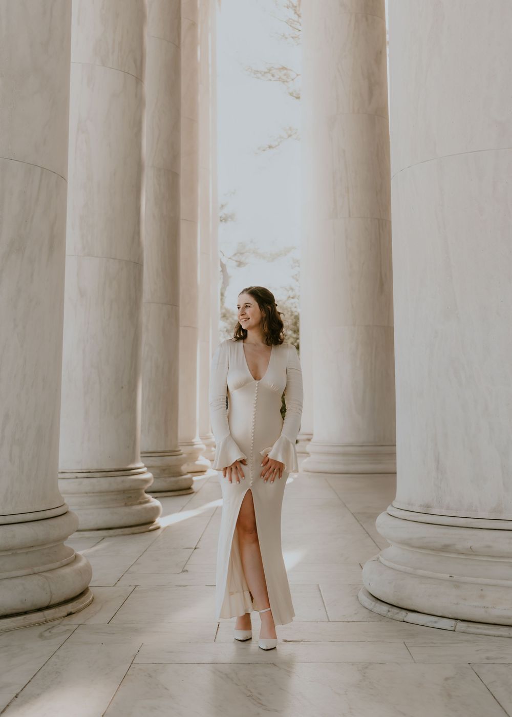 A person in a white dress stands elegantly between classical marble columns in a bright architectural setting.