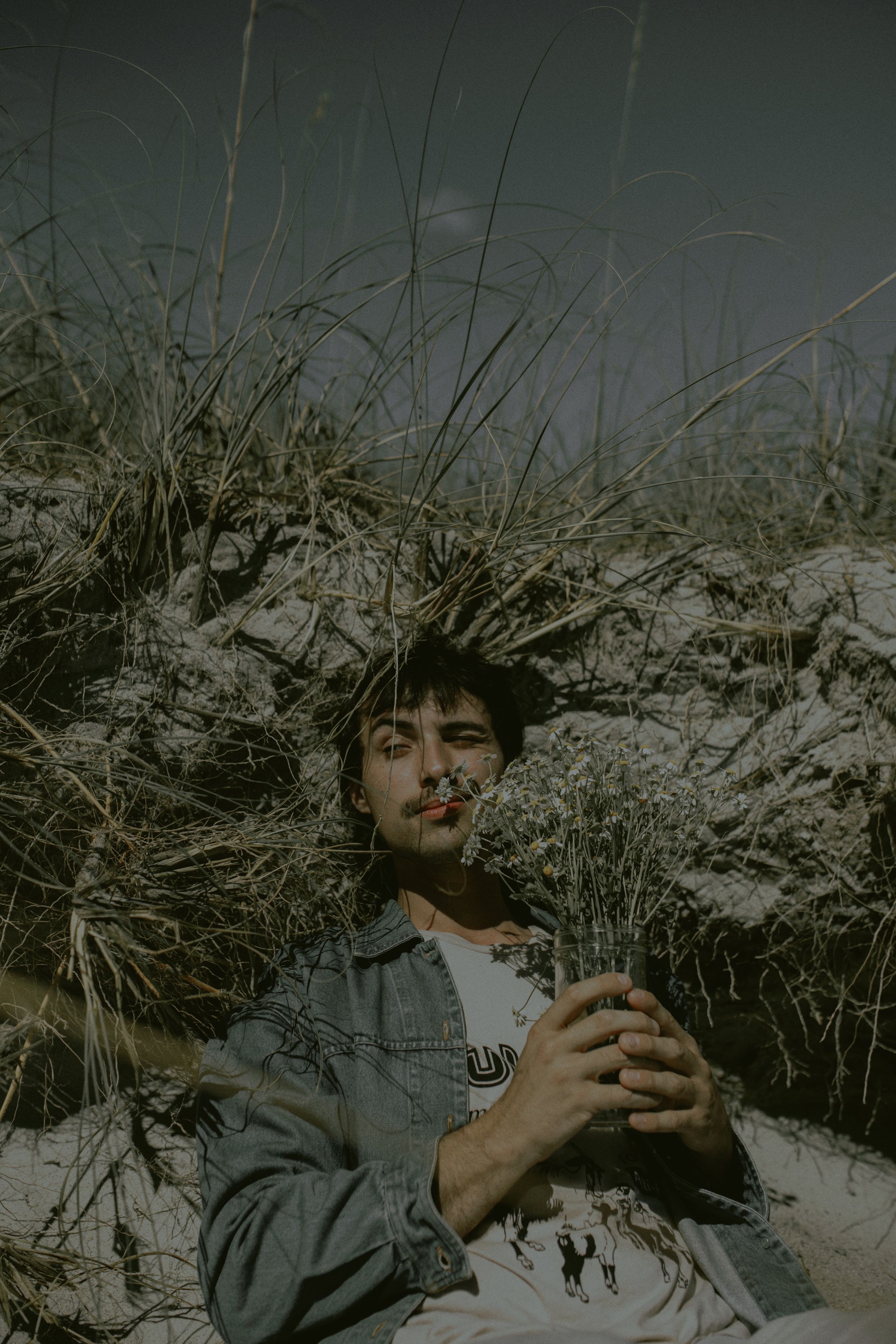 A person in a denim jacket lies in snow-covered grass while holding a warm beverage on a winter day.