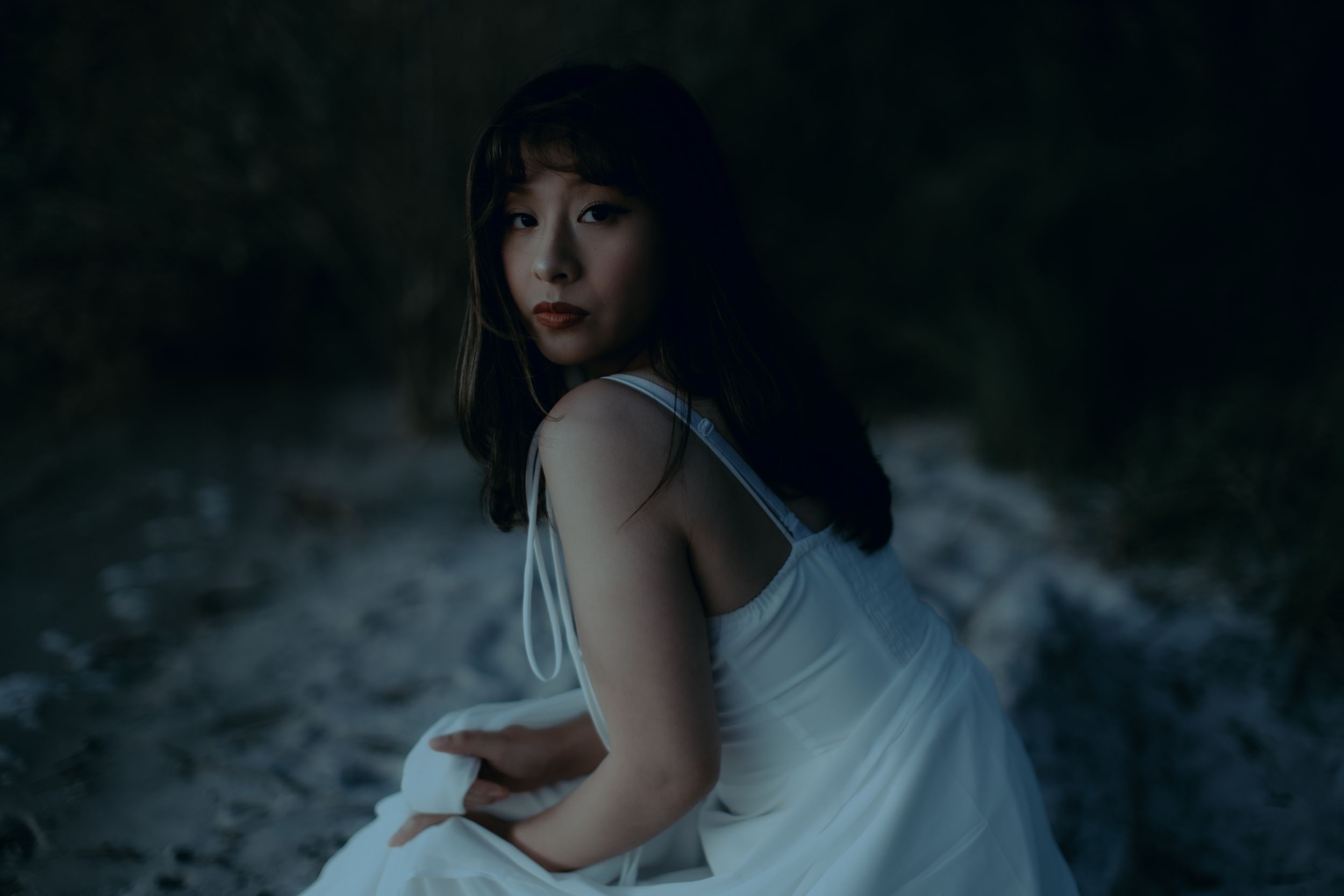 A person in a white dress poses against a dark background in a moody artistic photoshoot.