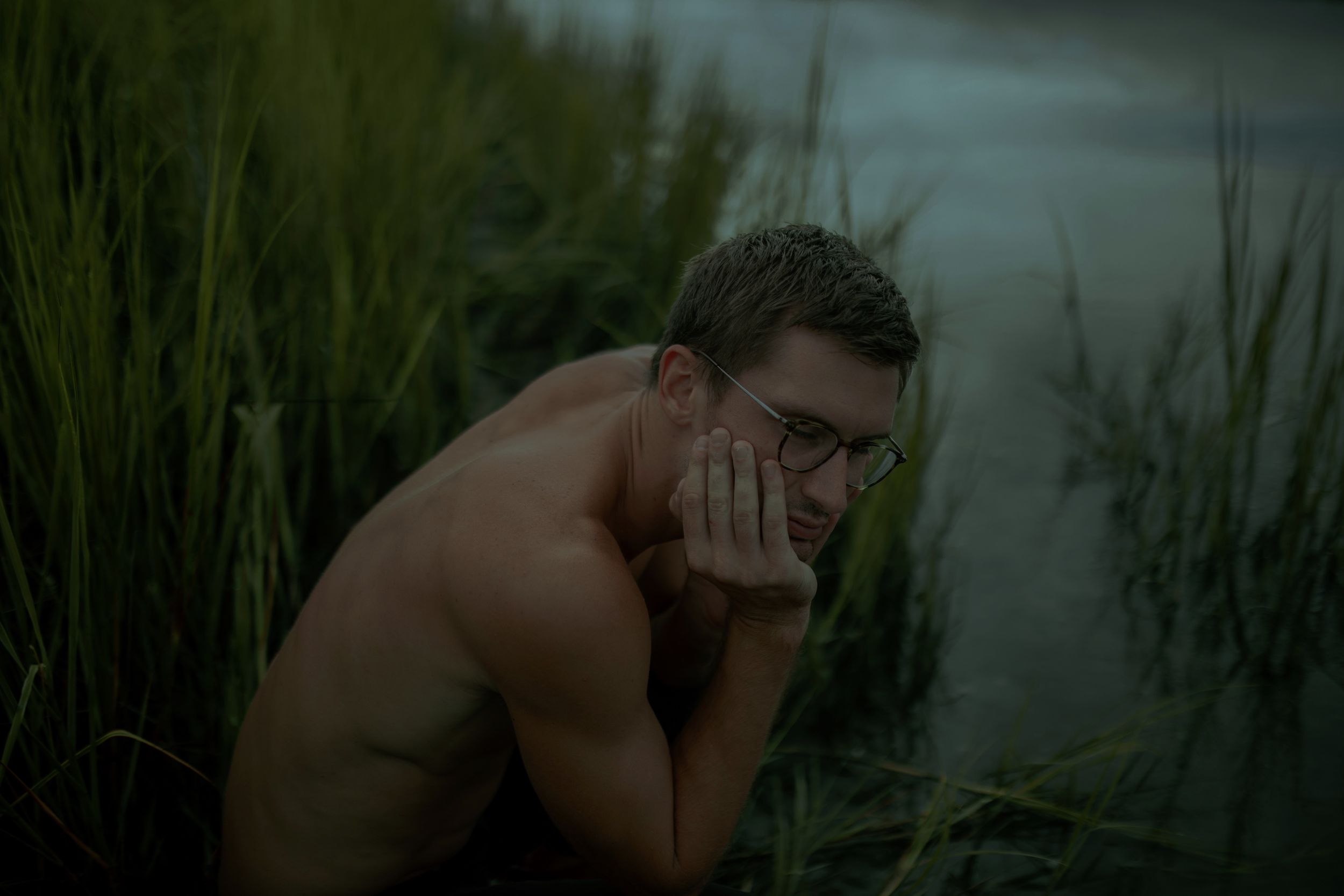A shirtless person sits thoughtfully in tall grass near water during golden hour lighting.