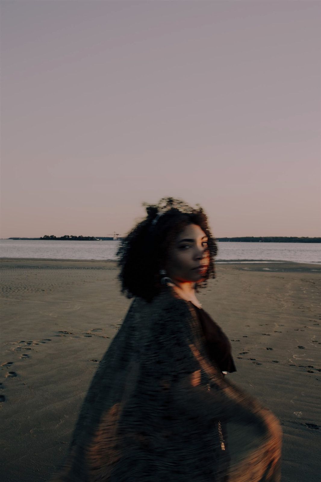 Silhouette of a person with flowing hair running along a beach at sunset against a soft pink sky.