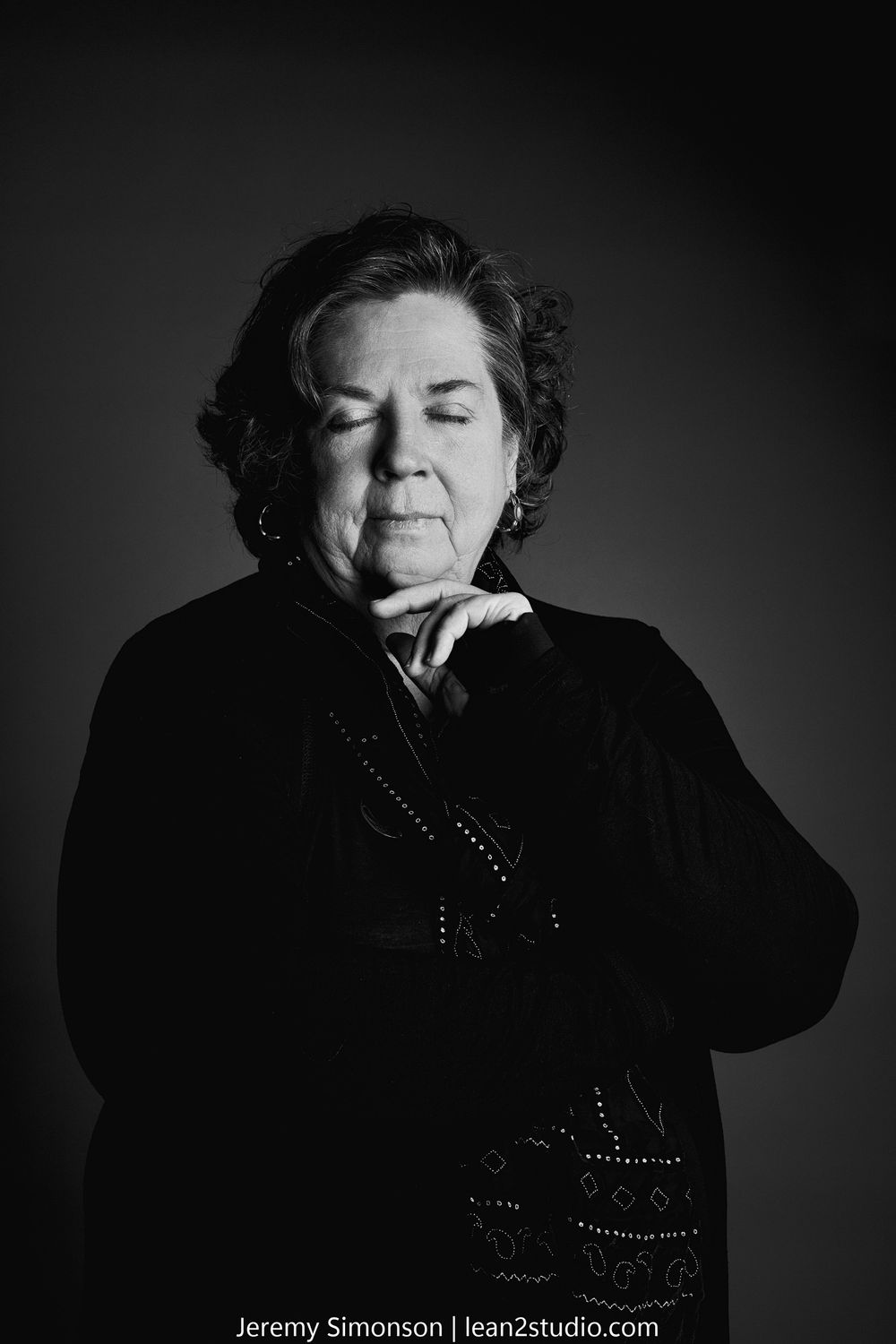 A dramatic black and white portrait of an elderly woman in dark clothing with closed eyes against a dark background.