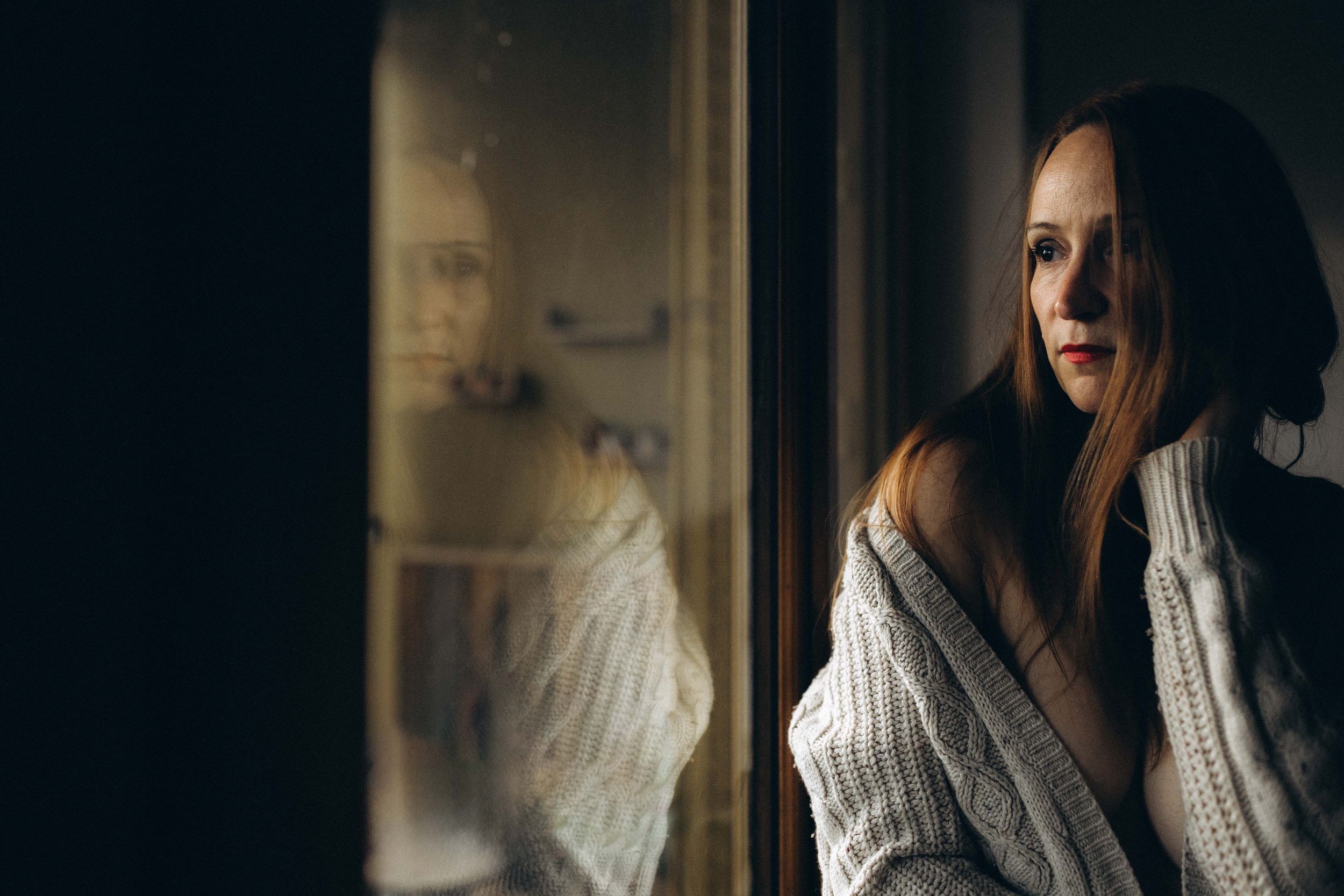 A person in a white knit sweater gazes contemplatively through a window with dramatic side lighting.
