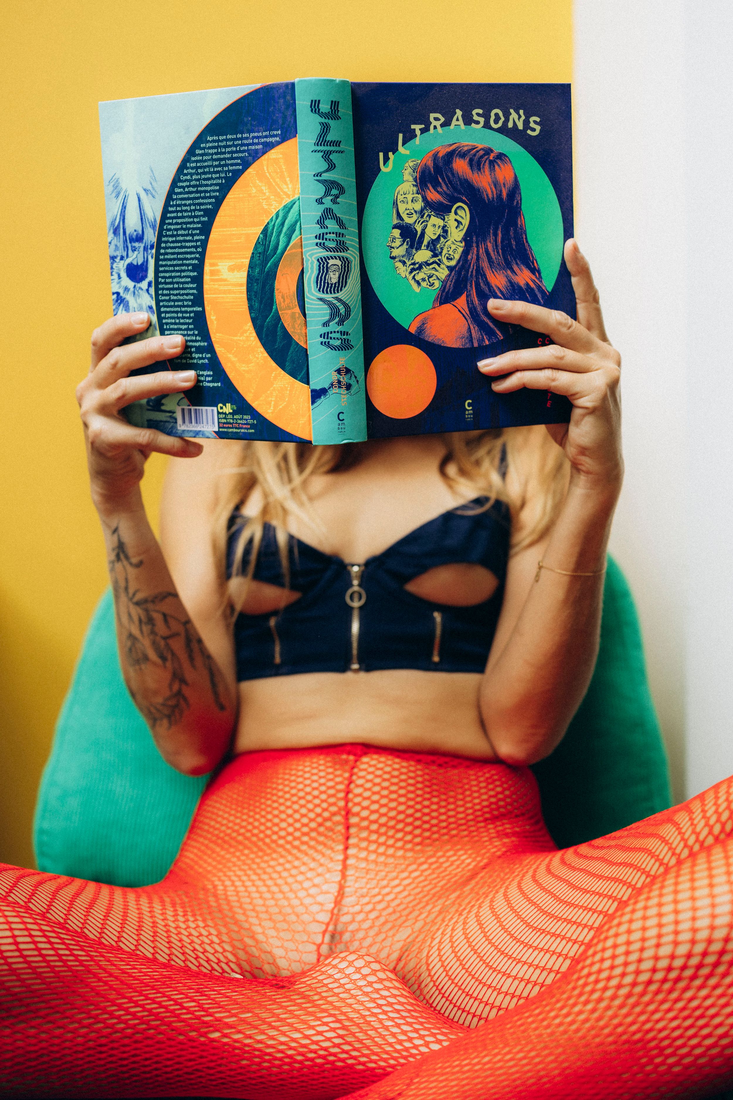 A person with colorful hair reads a book while wearing a green shirt and orange fishnet stockings.