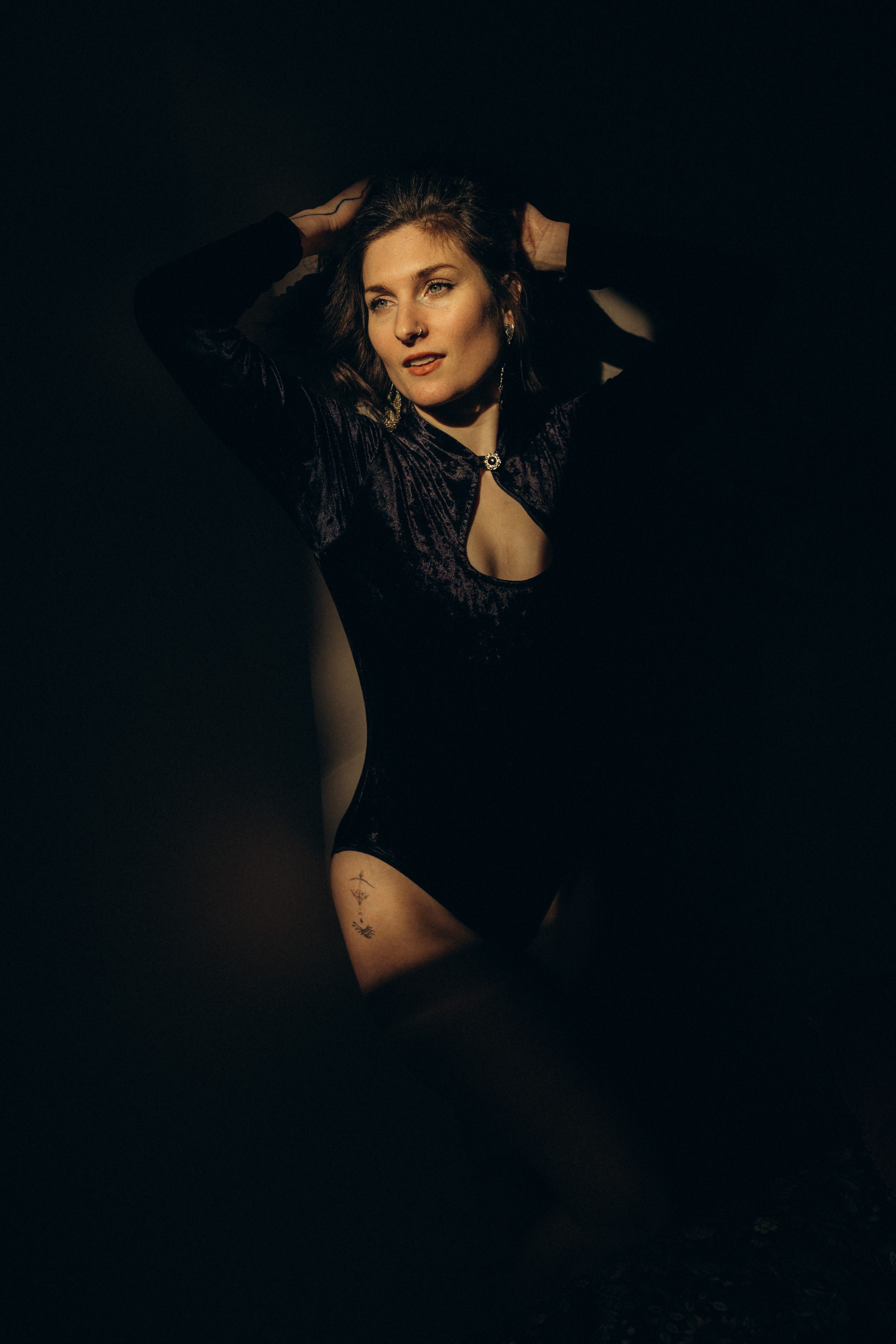 Moody boudoir portrait in black bodysuit against dark background with dramatic lighting and shadows.