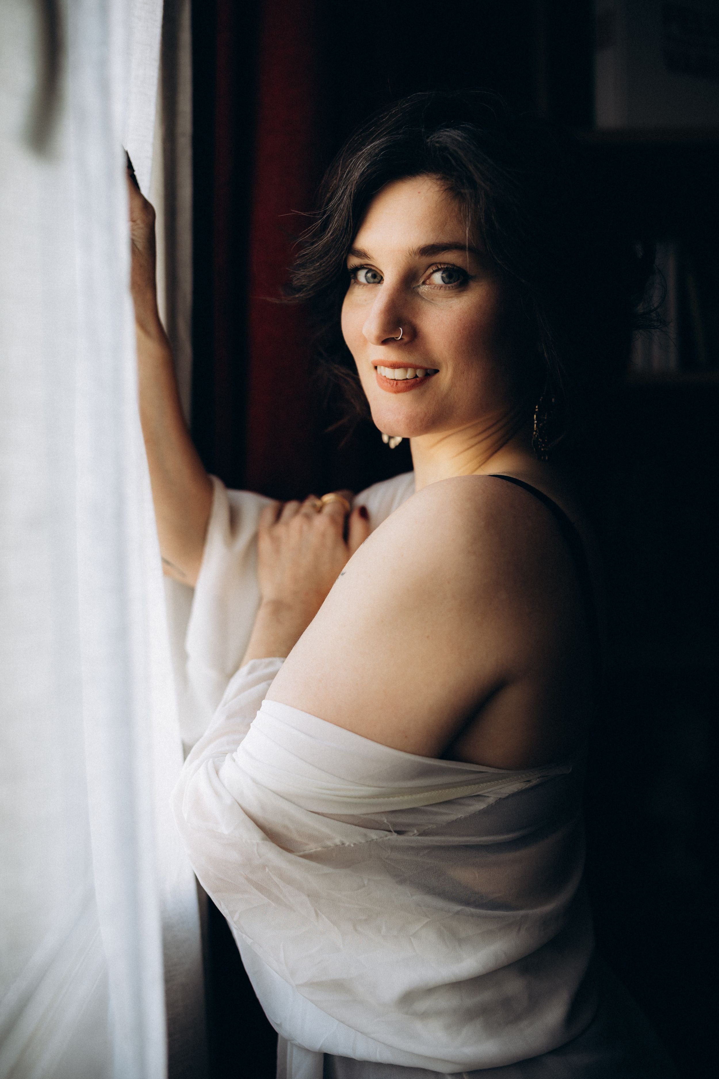 Portrait of someone in soft natural light by a window, wearing draped white fabric in an intimate boudoir style.