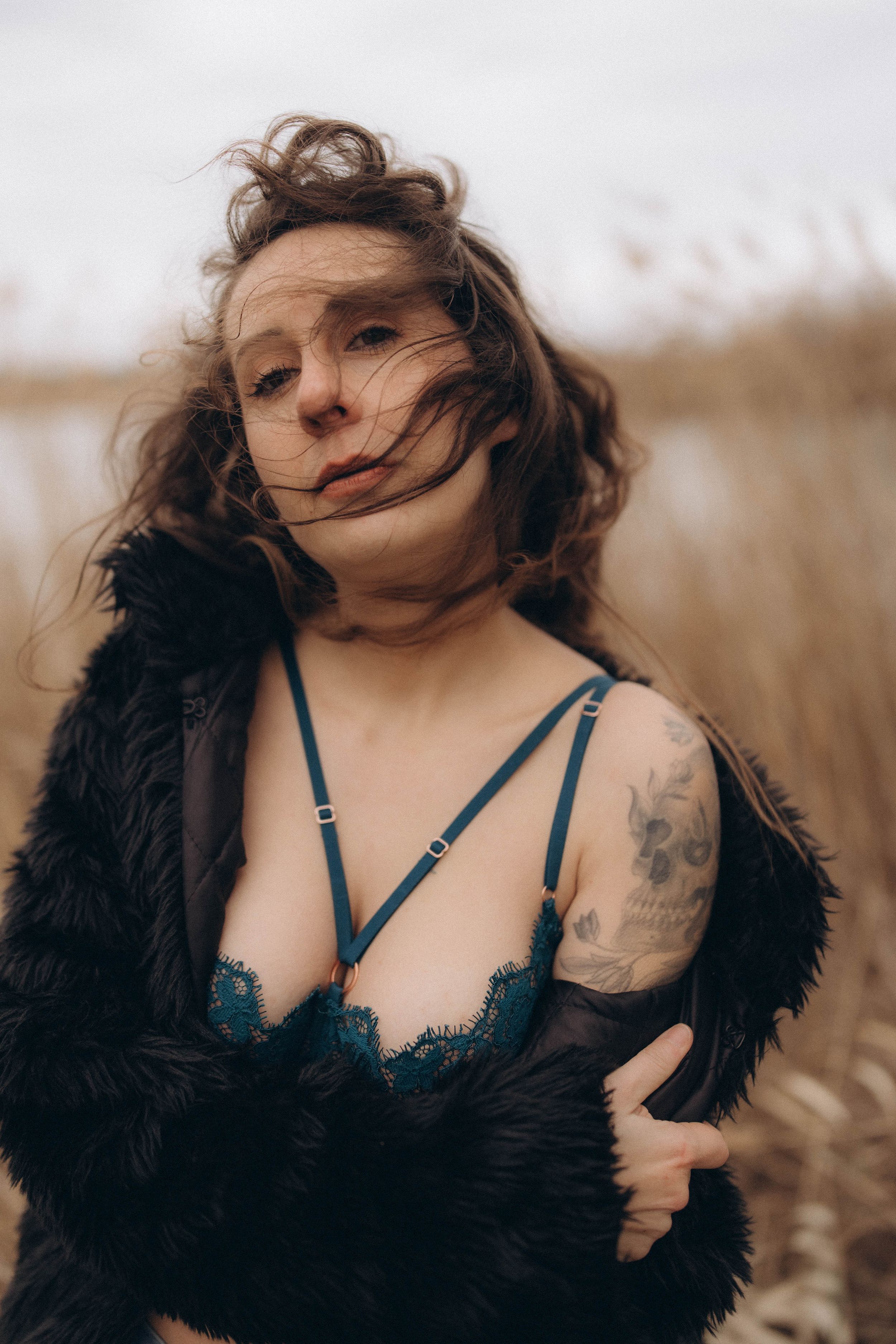 A figure in a black feathered coat and teal bra poses in a field with windswept hair in a moody photo series.