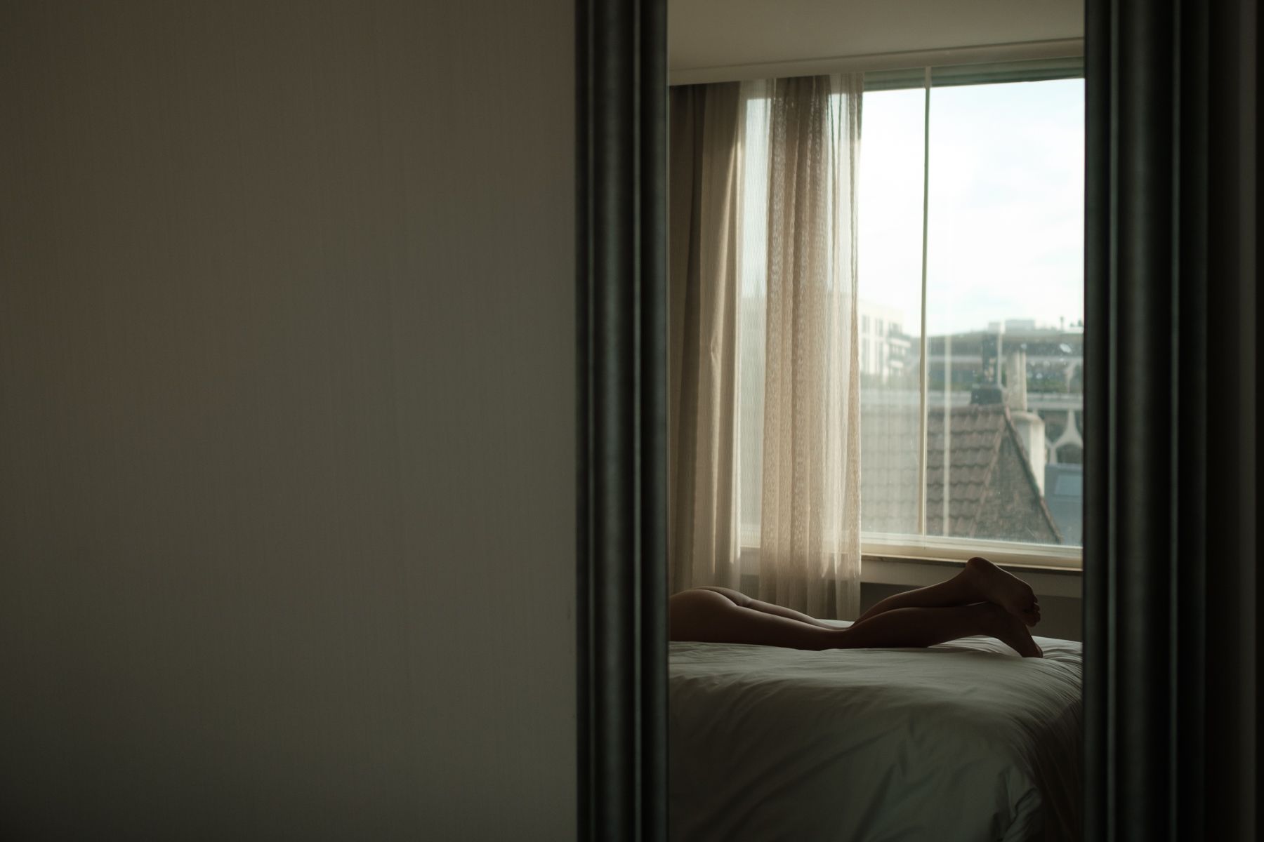 A dimly lit bedroom with sheer curtains and a window overlooking city buildings in the background.
