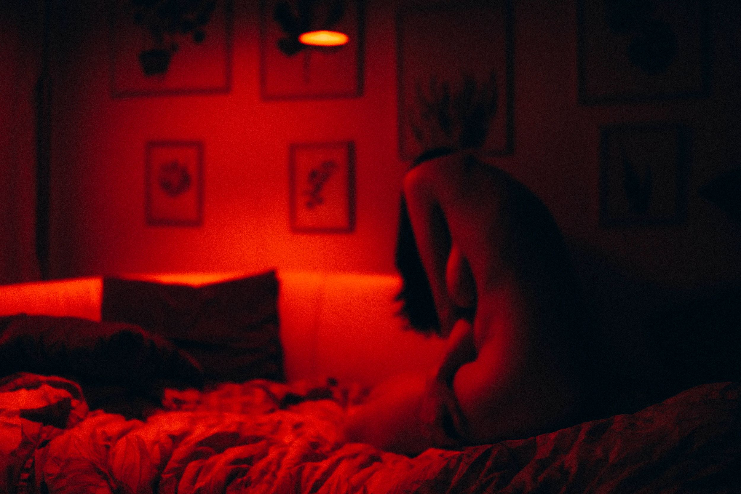 A silhouetted figure sits on a bed in a dimly lit room with red lighting and framed artwork on the walls.