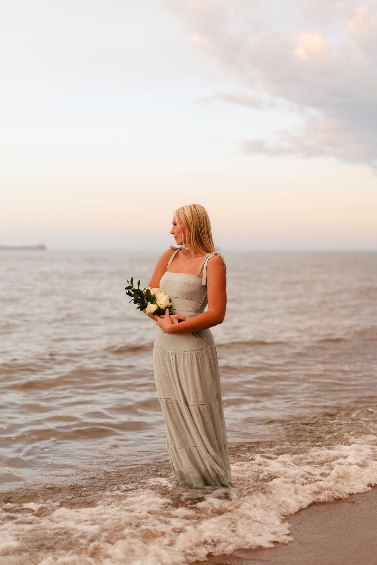 A figure in a flowing beige dress stands at the ocean's edge during sunset holding a bouquet of flowers.