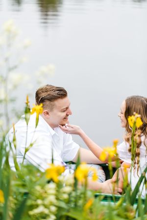 A couple sits together by a lake surrounded by blooming yellow flowers in a romantic outdoor setting.