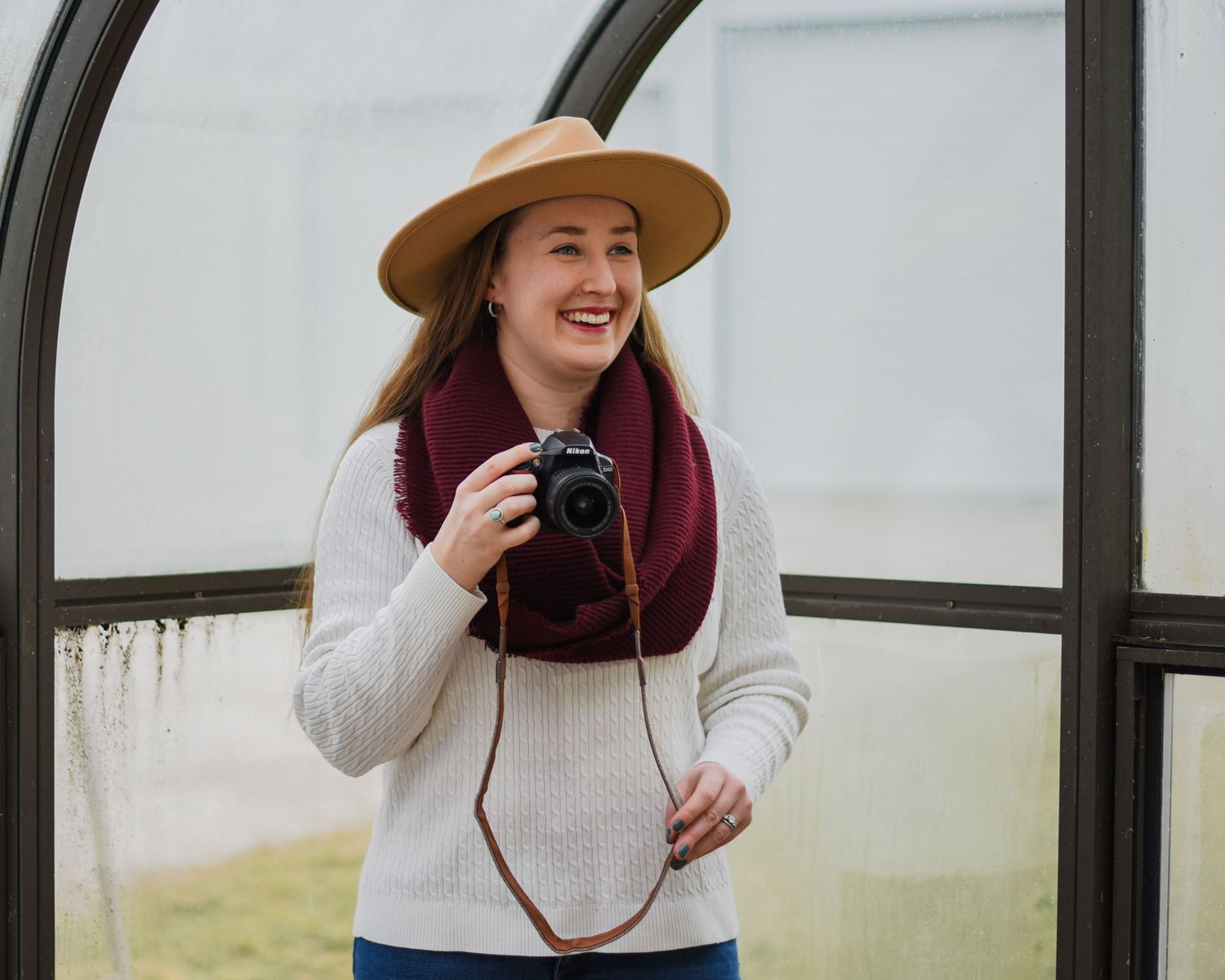 Person in white sweater and brown hat taking photos in a greenhouse setting.