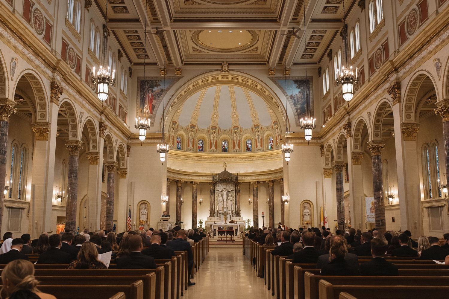 Interior view of an ornate Catholic church with arched columns, decorated ceiling and congregation seated in wooden pews.