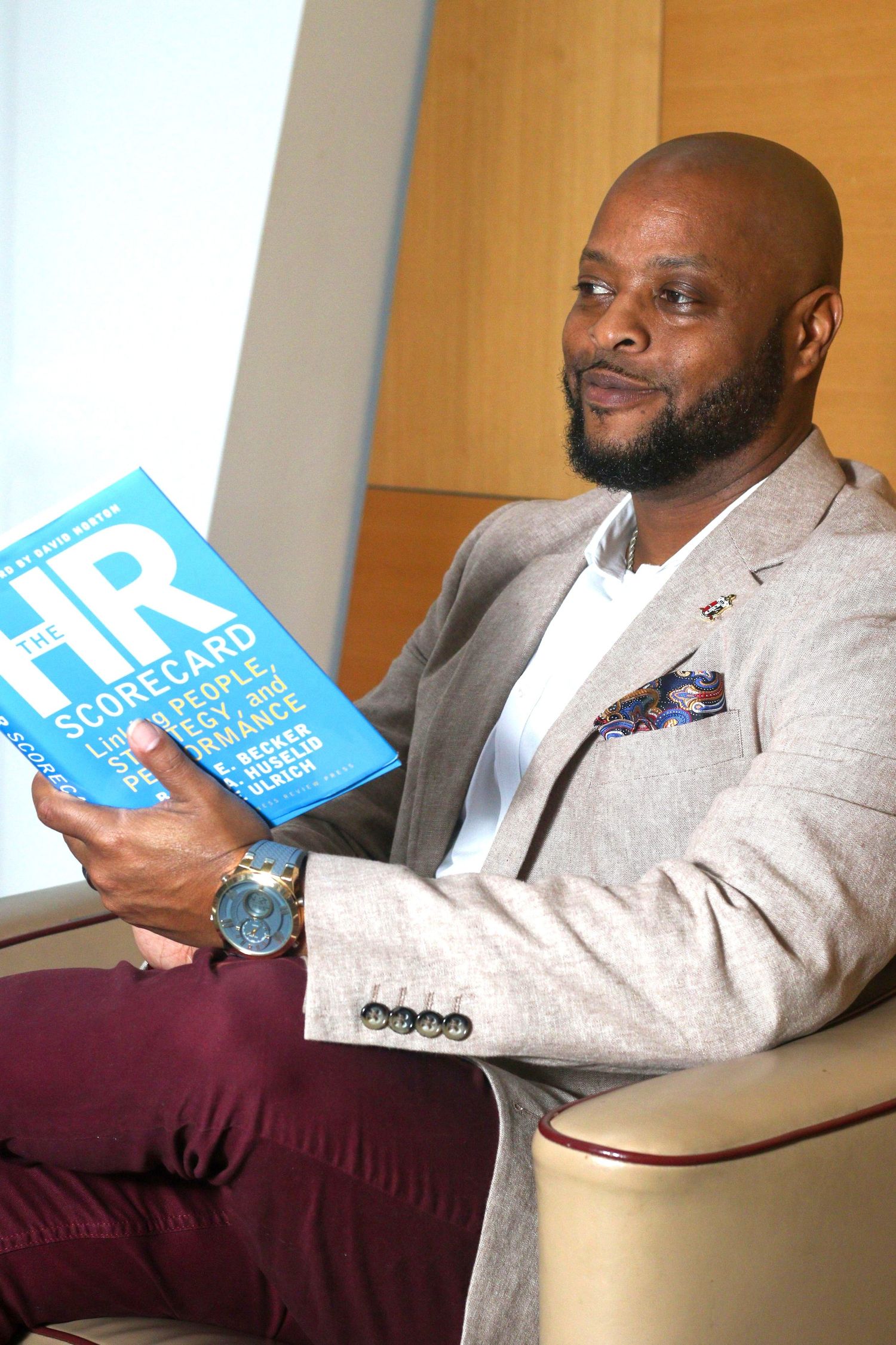 A person in a light blazer sits on a burgundy chair while holding a light blue book or document.