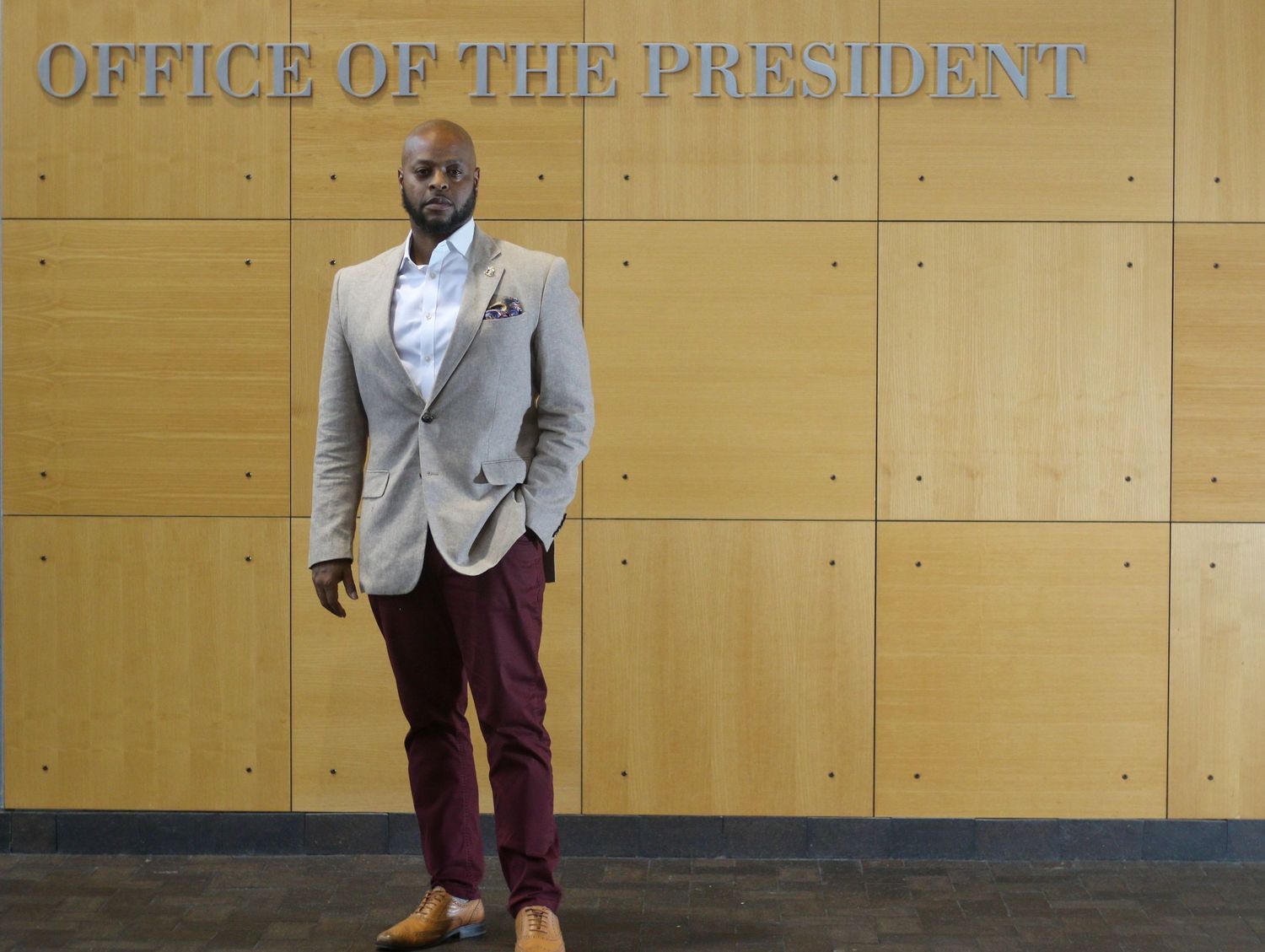 Person in a light gray blazer and burgundy pants stands in front of a wooden paneled wall with 'Office of the President' text.