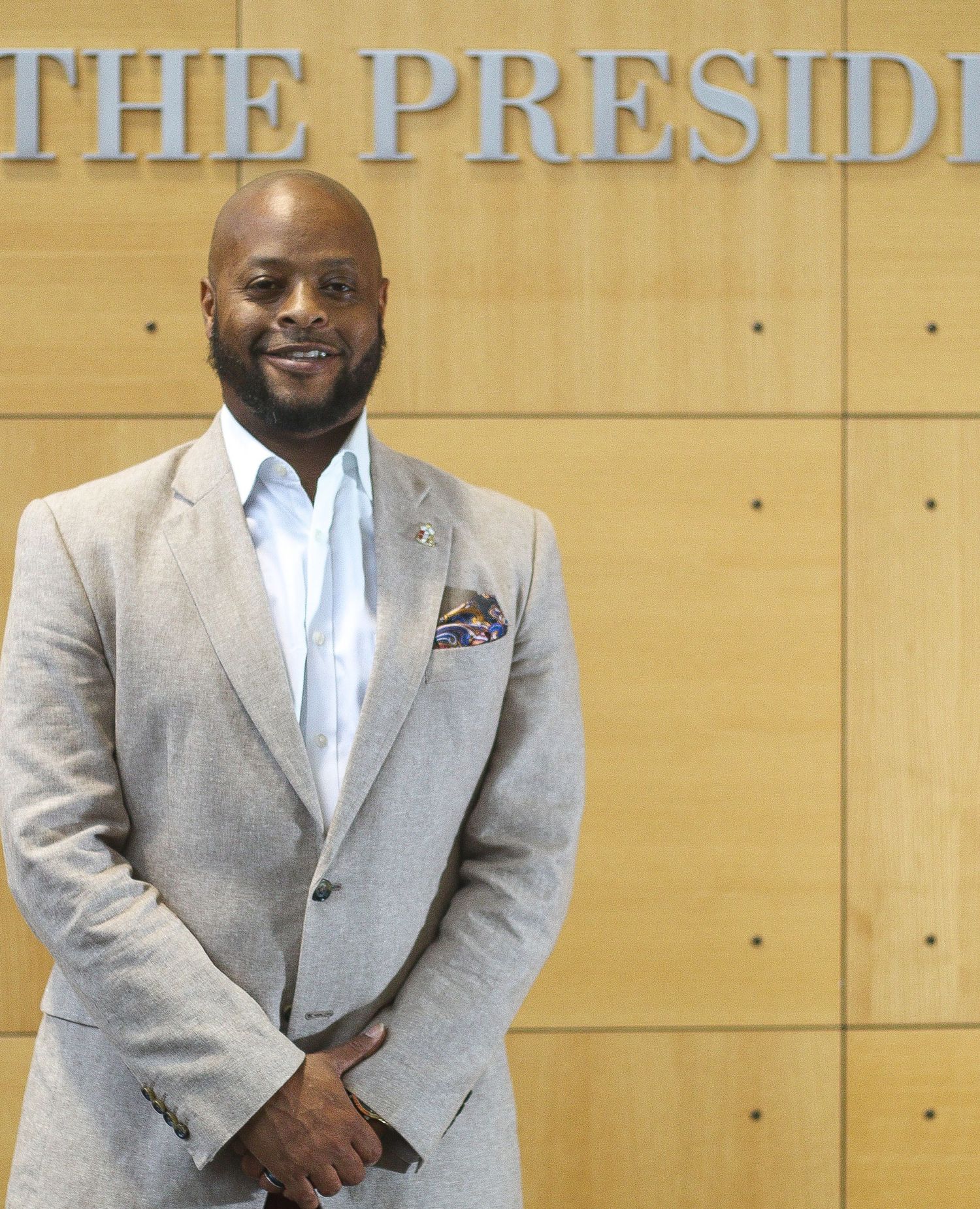 Professional wearing a light gray suit and white dress shirt poses against a wood-paneled wall with text signage.