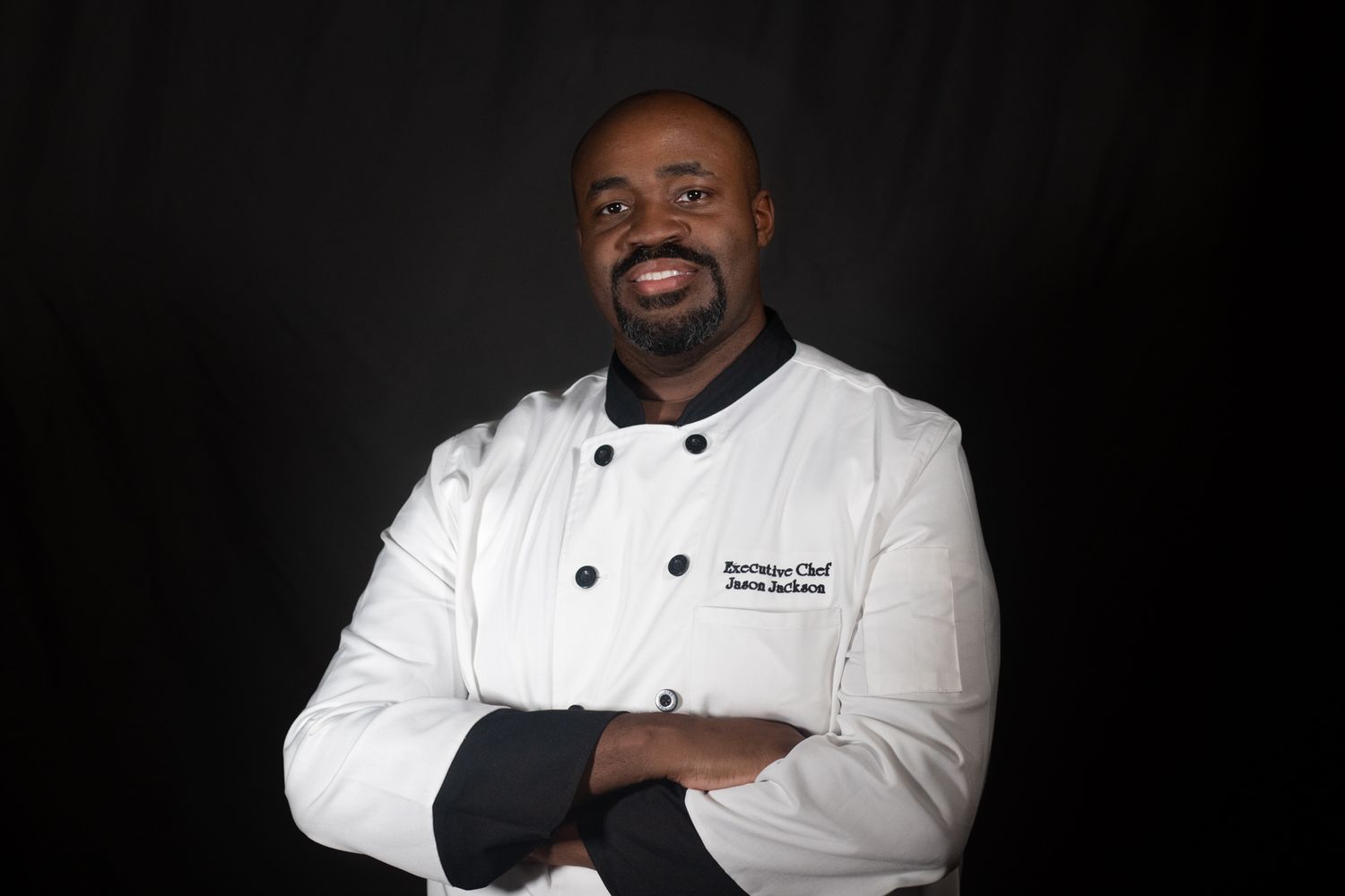 A professional chef poses in white uniform against dark background.