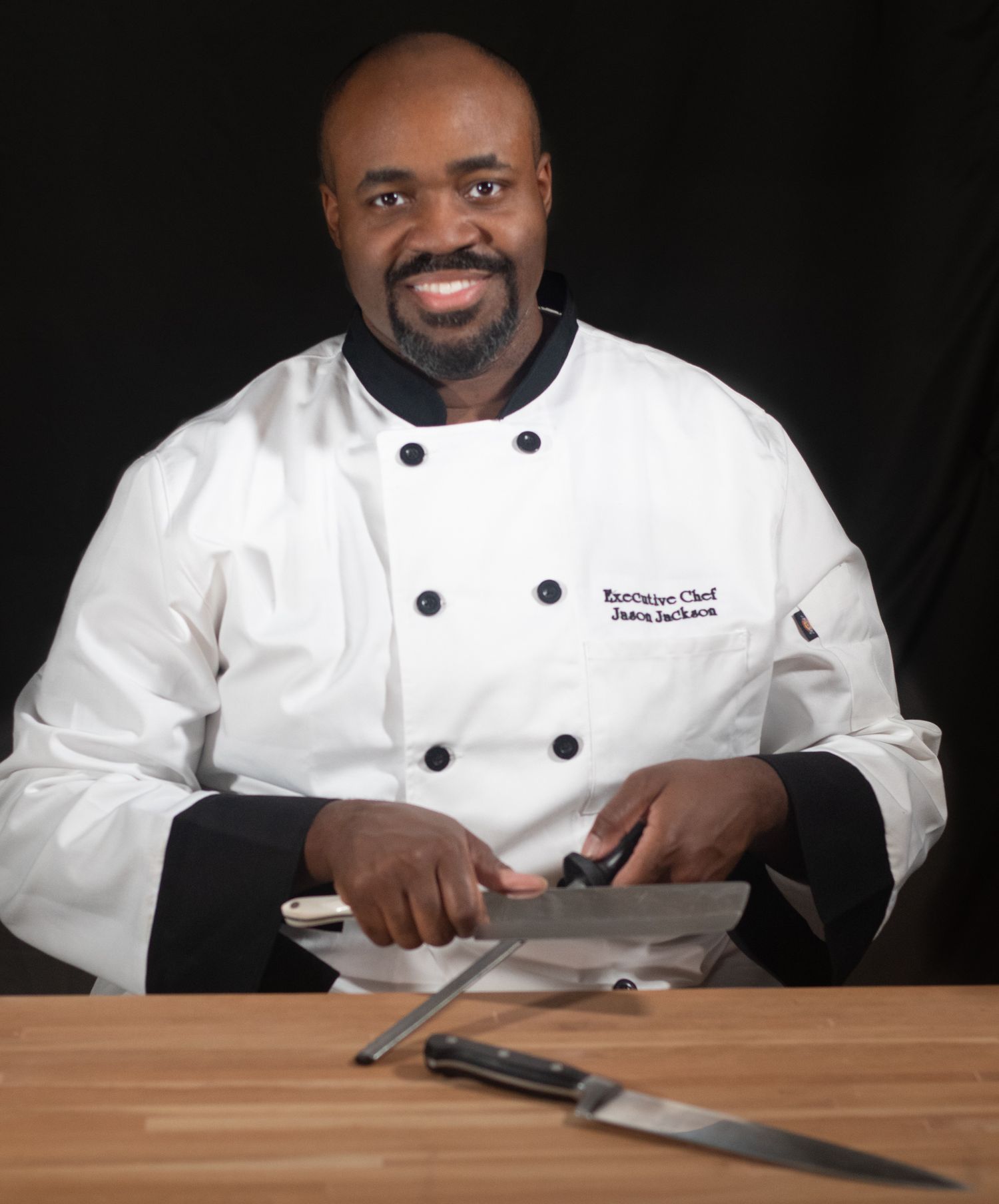 A chef in white uniform sharpens a knife while sitting at a wooden table in a professional kitchen setting.