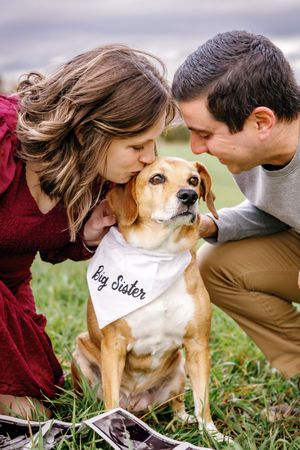 A couple cuddles with their dog wearing a bandana in an outdoor setting.