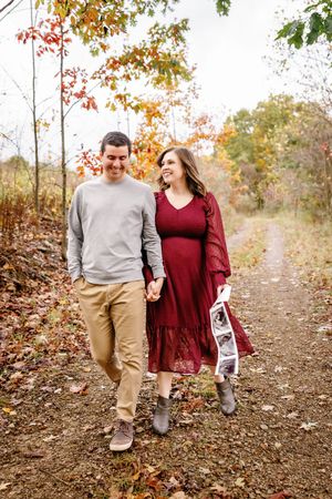 A couple walks hand in hand on a leaf-covered path during fall, holding ultrasound photos.
