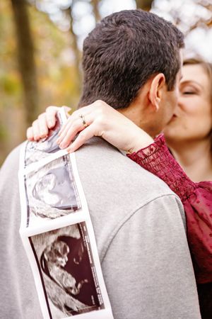 A couple holds up ultrasound photos while embracing during an autumn pregnancy announcement photoshoot.