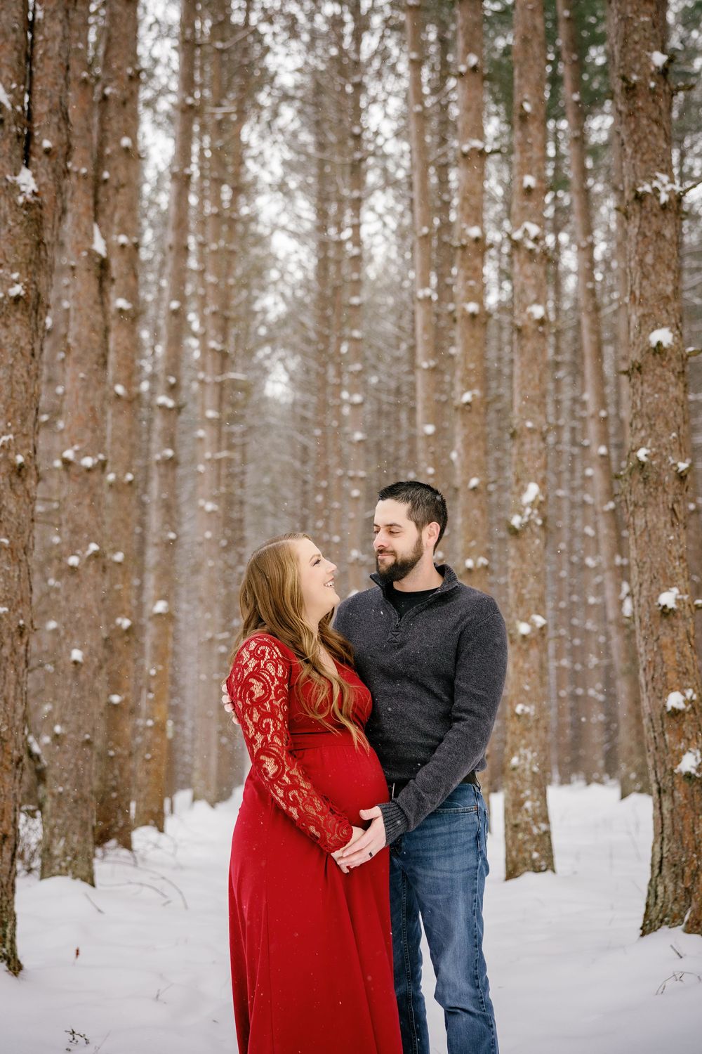 A couple shares an intimate moment in a snowy forest with tall pine trees.