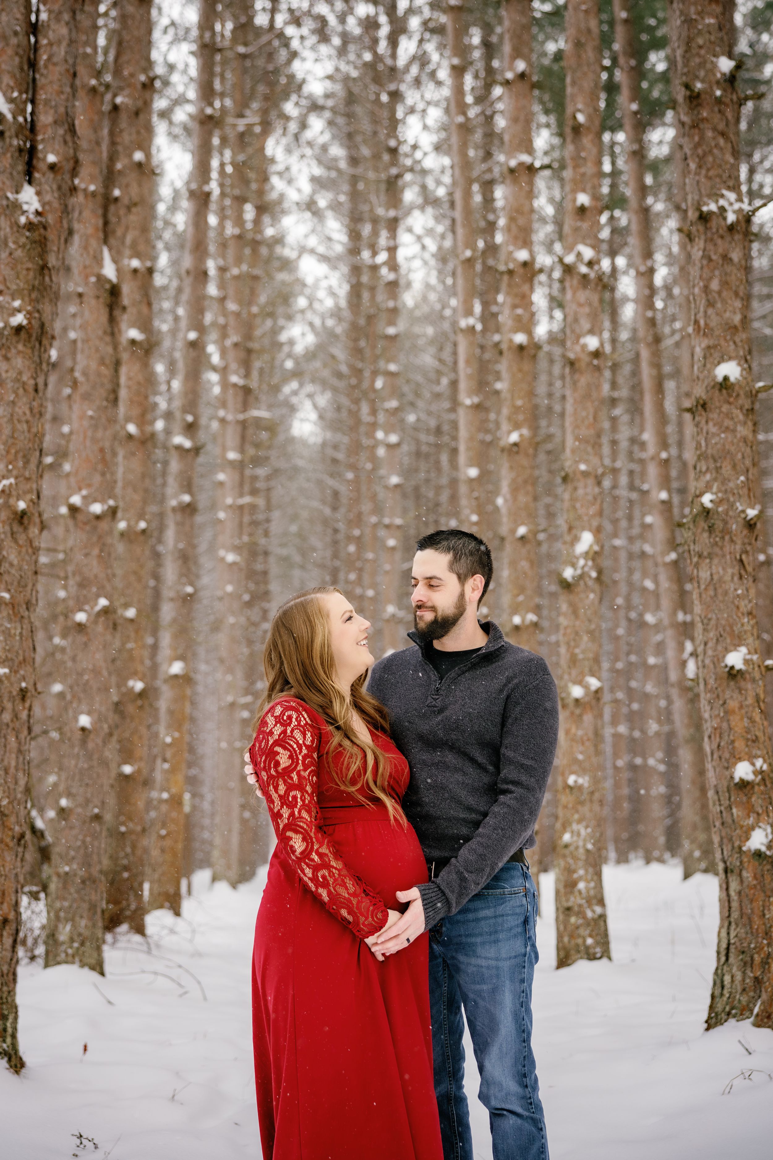 A couple shares an intimate moment in a snowy forest with tall pine trees.