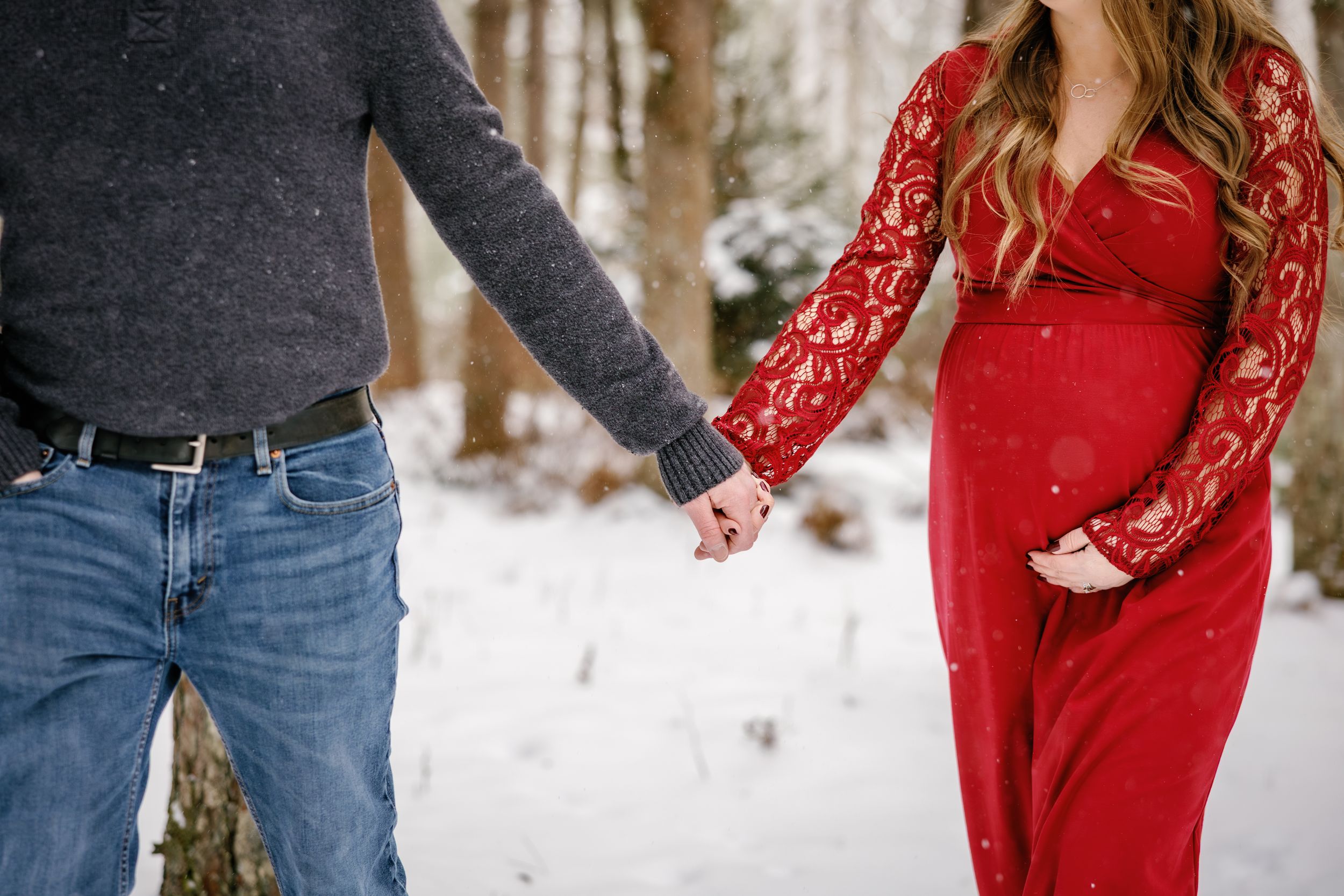 Close-up of holding hands in winter, with red lace dress against snowy forest background.