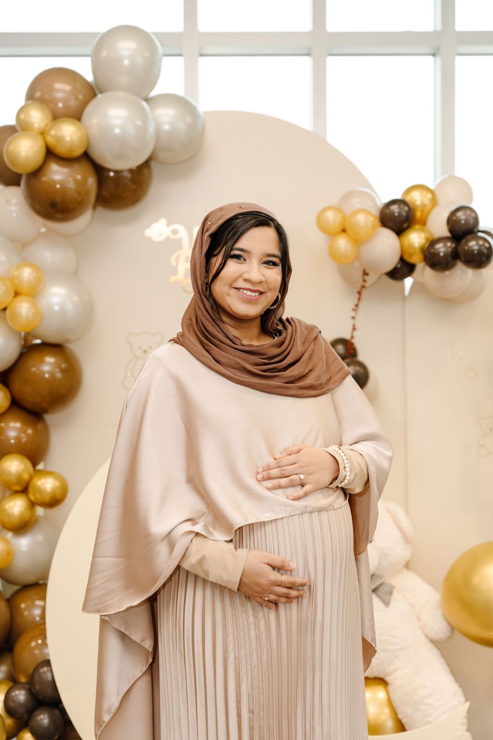 Smiling pregnant woman in beige hijab and pleated dress poses at gold and white balloon baby shower backdrop.