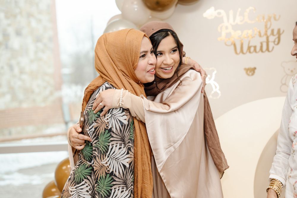 Two women in hijabs share a warm embrace at an elegant gold and white baby shower celebration.