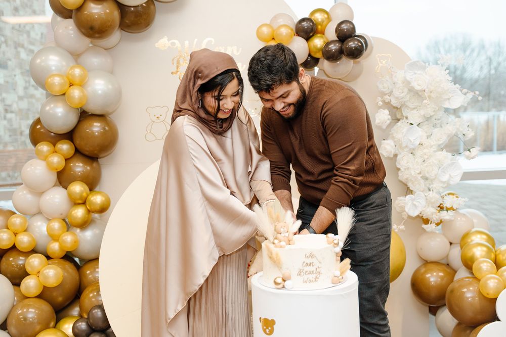 Muslim couple cutting white baby shower cake together surrounded by gold and brown balloon arch decor.