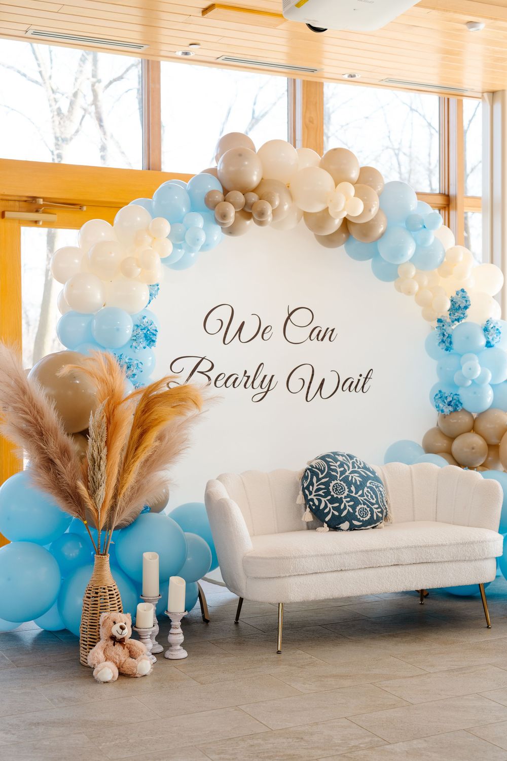 A elegant baby shower backdrop with blue and gold balloon arch and 'We Can Bearly Wait' sign above a white sofa.