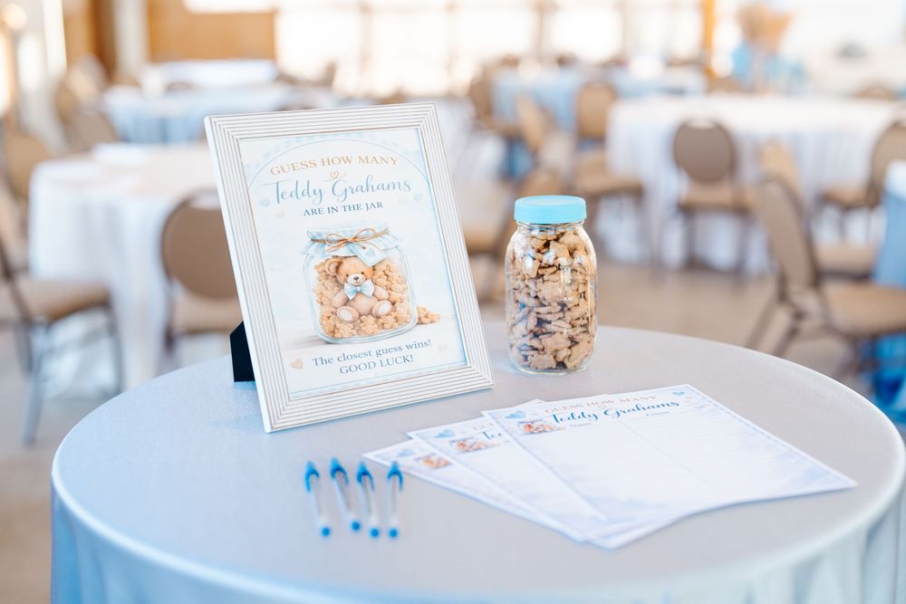 Baby shower teddy graham guessing game with jar, sign, and blue pens on a light blue tablecloth.