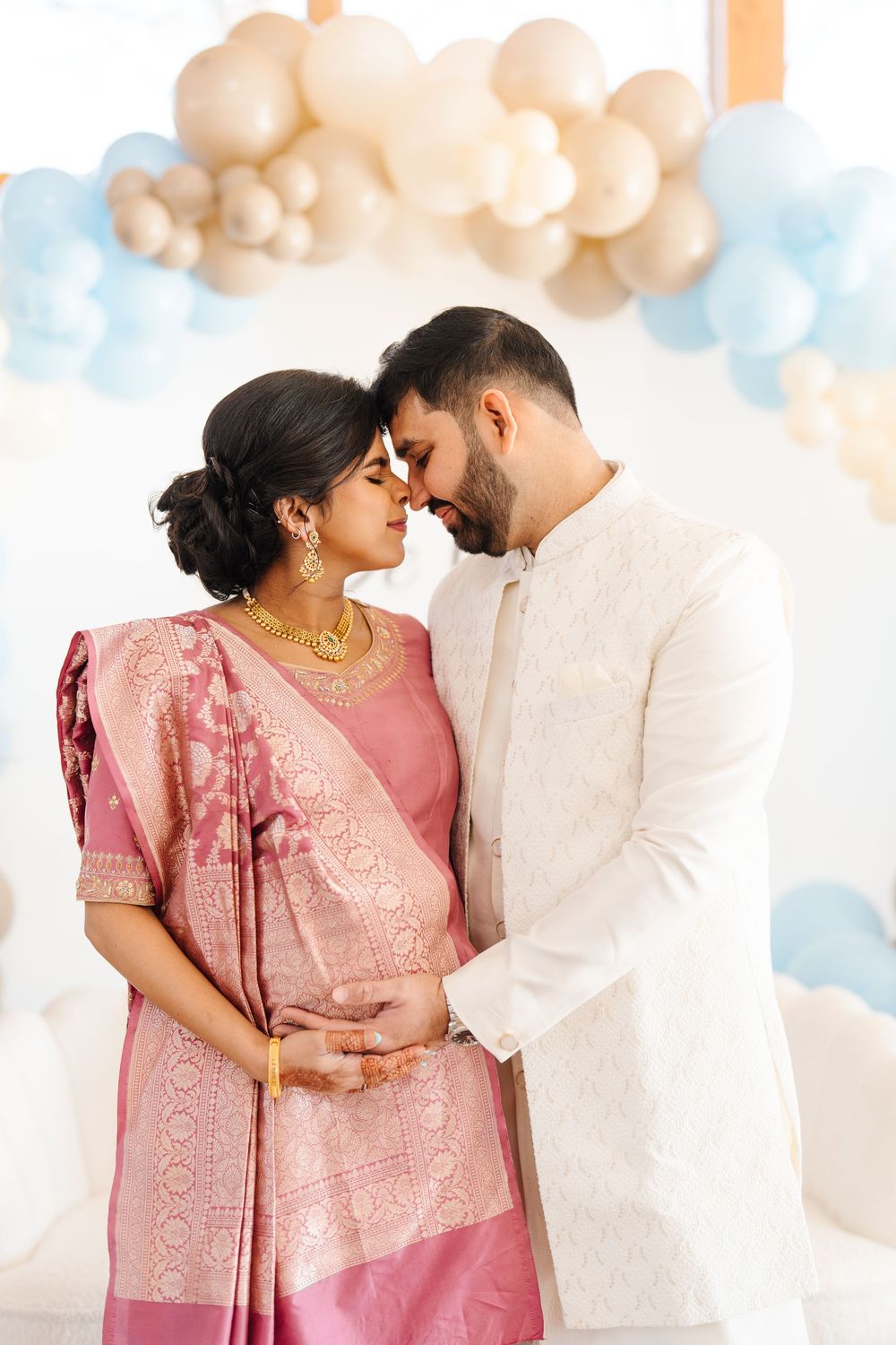 Pregnant Indian couple in traditional attire tenderly touching foreheads at a blue and beige baby shower.