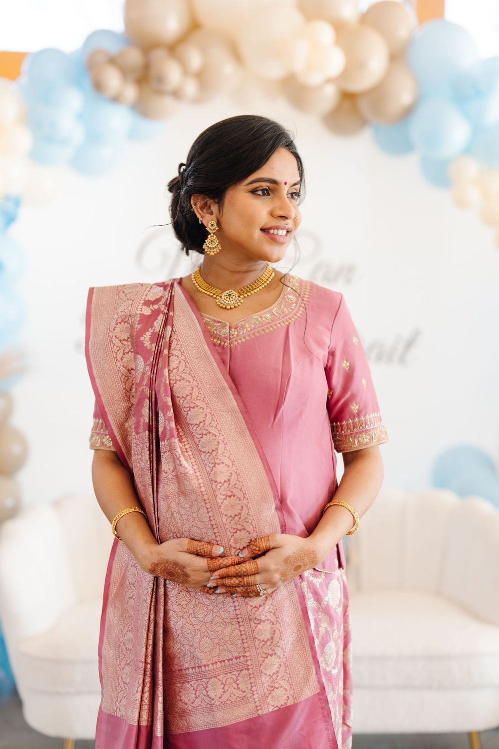 Smiling pregnant Indian woman in pink silk saree with gold jewelry at a baby shower celebration.