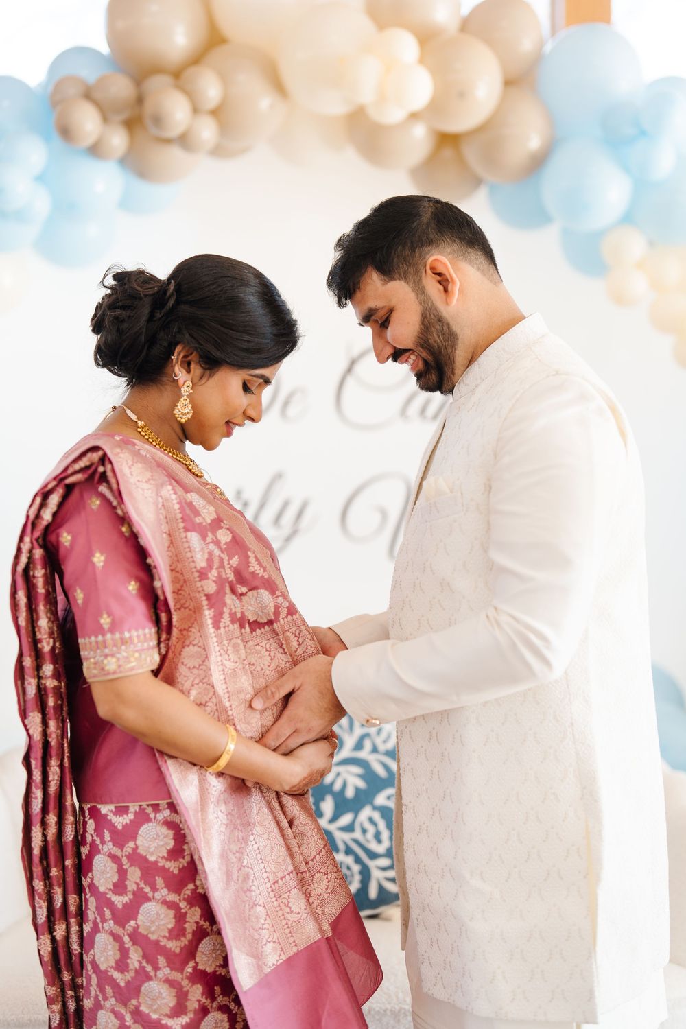 Indian couple in traditional attire lovingly looking at pregnant belly at a blue balloon decorated shower.